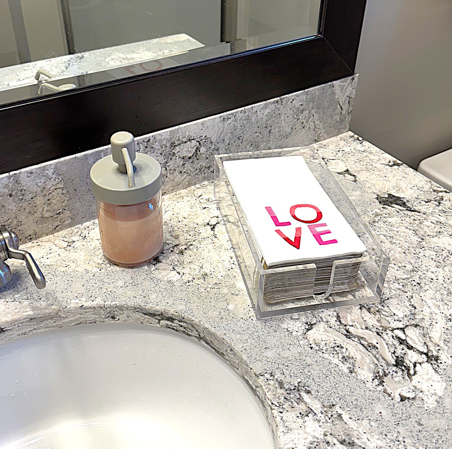 A clear acrylic napkin holder with novelty napkins placed in bathroom.