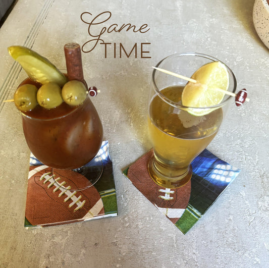 Two cocktail glasses with appetizer skewers; one glass contains a Bloody Mary cocktail, and the other has a beer-like drink. Both glasses are adorned with football-themed napkins.