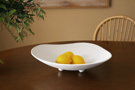 A white melamine bowl with a wavy, organic shape and small feet, shown on a dark wooden table with three yellow lemons inside. A wooden chair and a beige wall are visible in the background.