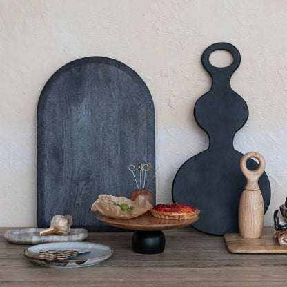 Two black cutting boards with handles, brown and black pedestal serving piece on a wooden surface against a light wall.