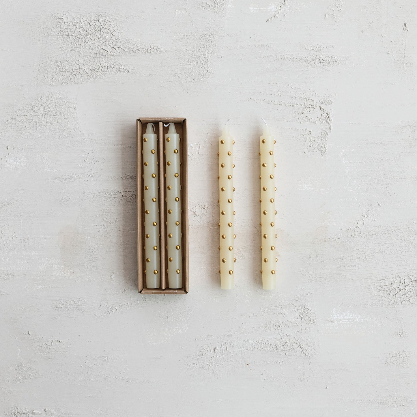 Three cream taper candles with small gold dots, with two inside a brown cardboard box 