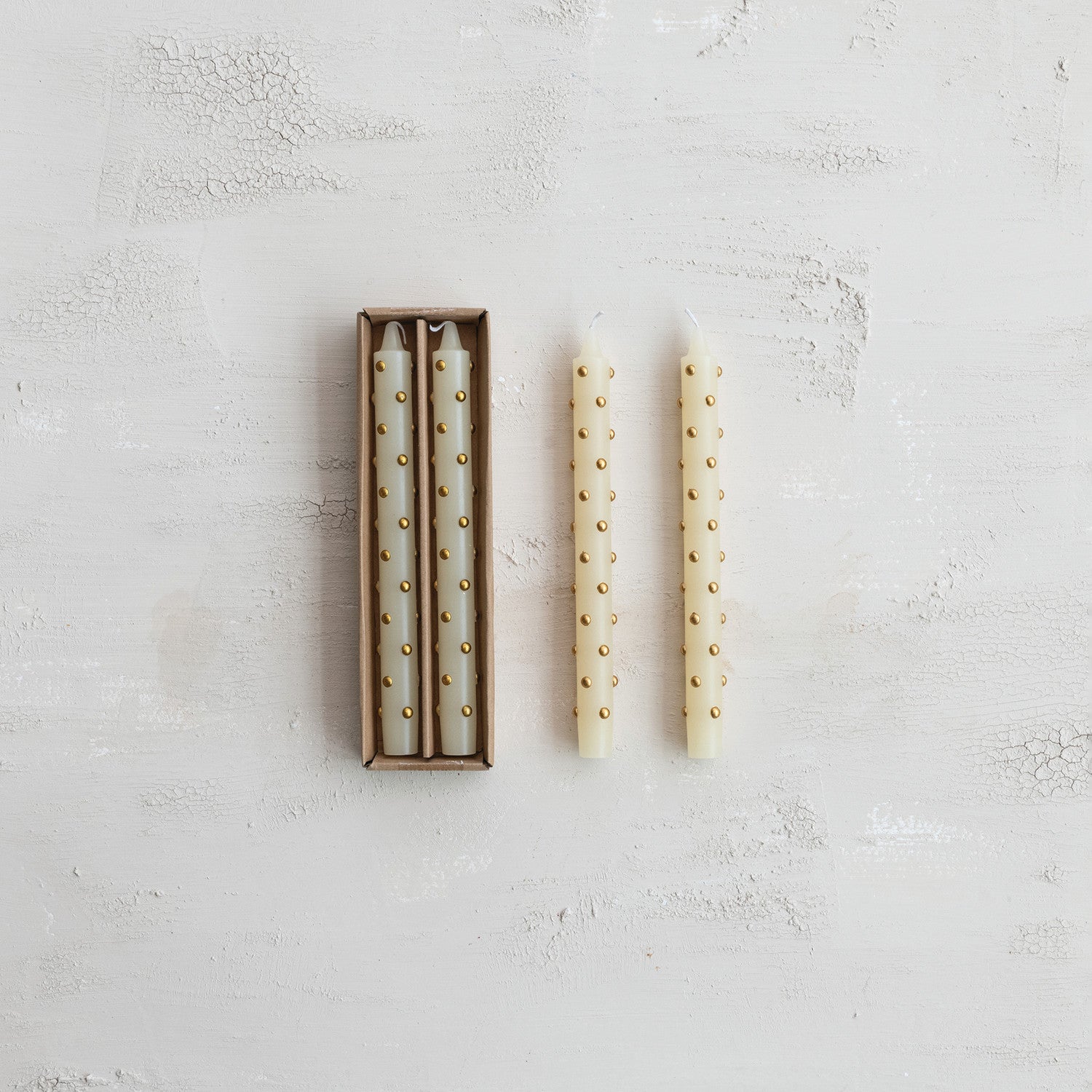 Three cream taper candles with small gold dots, with two inside a brown cardboard box 