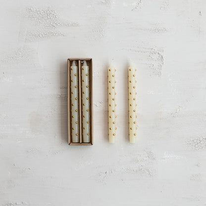 Three cream taper candles with small gold dots, with two inside a brown cardboard box 