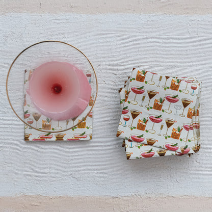 A set of cocktail-themed paper napkins with a white background and colorful printed drinks, shown with one napkin folded under a clear glass coaster and a stack of folded napkins on a textured white surface.